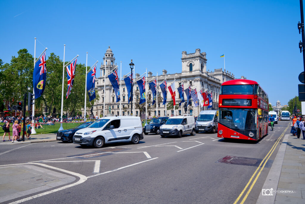 ulica Londynu z czerwonym, piętrowym autobusem. W tle flagi Anglii oraz budynek Royal Institution of Chartered Surveyors. Fotoskierka sees a London street with a red double-decker bus. In the background are the flags and the Royal Institution of Chartered Surveyors.