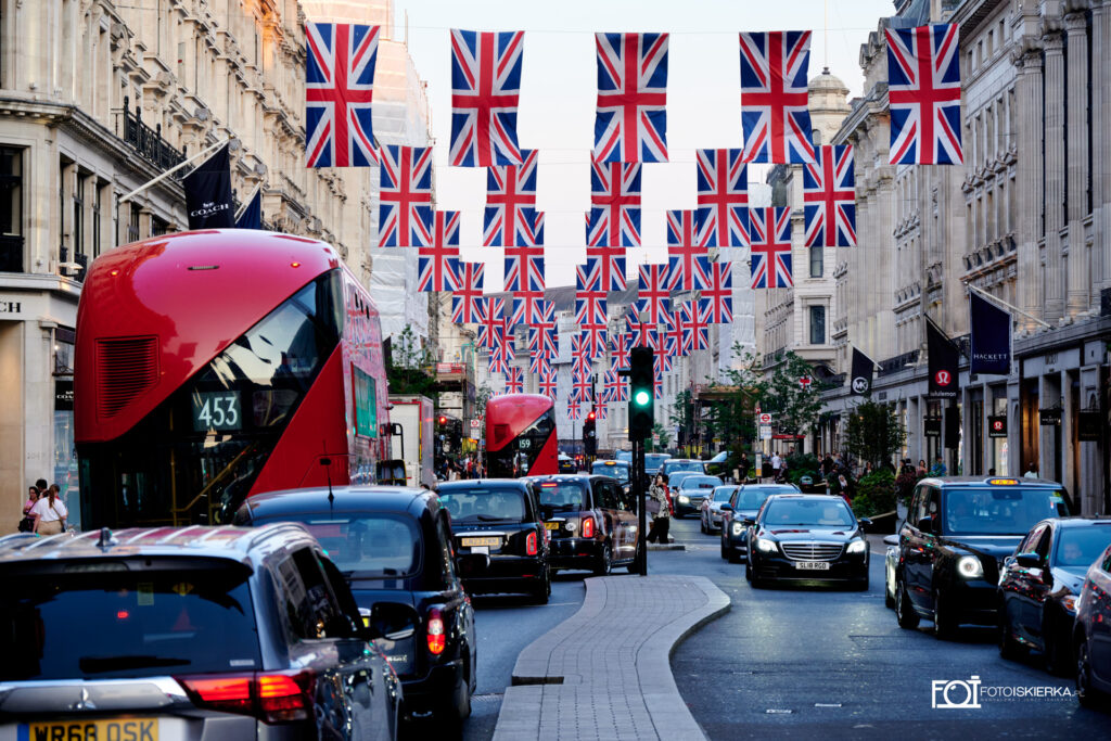 ulica w Londynie w Anglii z flagami, czerwonym autobusem i samochodami. The photo spark sees a street in London, England with flags, a red bus and cars.