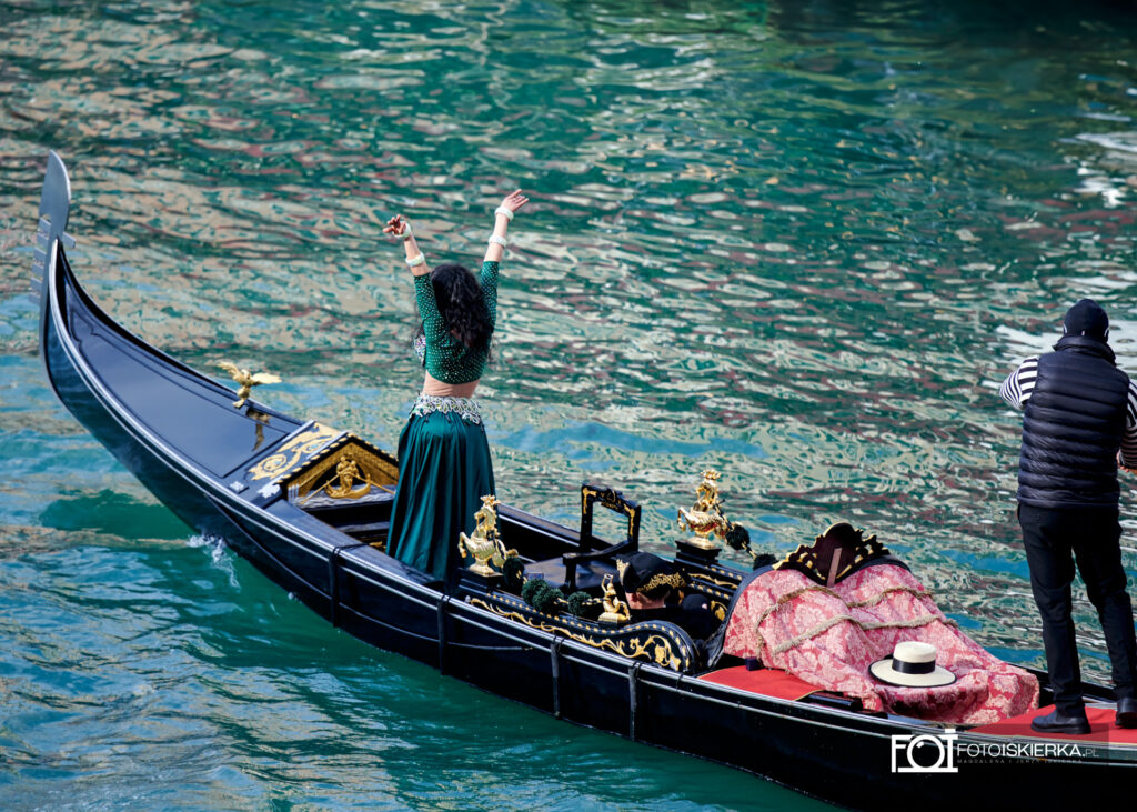 Zbliżenie z góry na czarną gondolę płynącą po turkusowej wodzie. Na pokładzie kobieta w szmaragdowym stroju do tańca brzucha wznosi ręce w geście radości. Obok niej stoi mężczyzna w pasiastej koszulce, a tył łodzi zdobią złote figury koników morskich i czerwona wzorzysta tkanina.