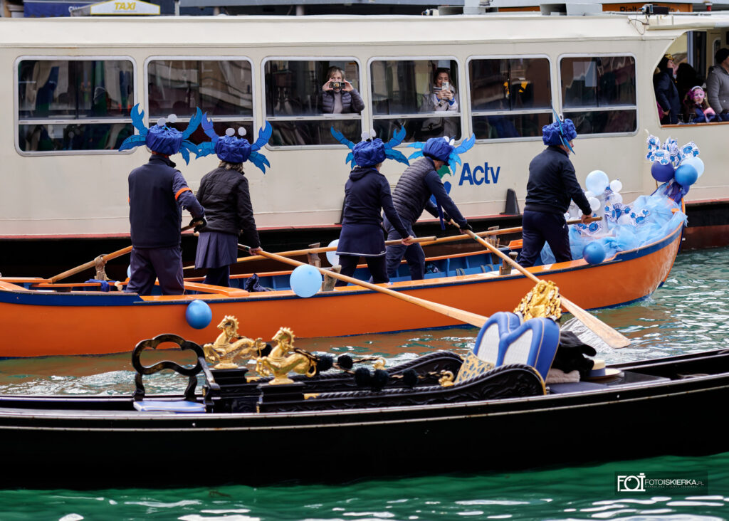Pomarańczowa łódź wiosłowa na Kanale Grande, której załoga ma na głowach duże, niebieskie czapki w kształcie krabów. W tle widać biały tramwaj wodny (vaporetto) z pasażerami obserwującymi paradę.