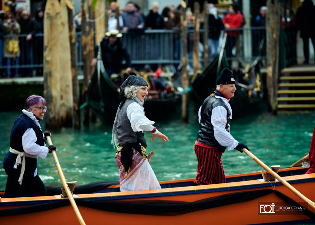 Trzy osoby w strojach pirackich i ludowych wiosłują w pomarańczowej łodzi. Na pierwszym planie uśmiechnięta kobieta w białej koszuli z falbanami i ciemnej kamizelce. W tle widać tłum gapiów na nabrzeżu oraz cumujące gondole.