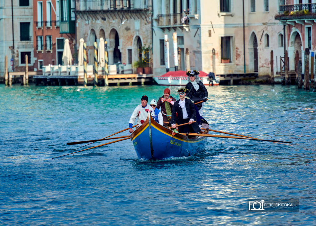 Błękitna łódź płynąca środkiem kanału, obsadzona przez wioślarzy w historycznych i fantazyjnych kostiumach. Jeden z mężczyzn ma na sobie białą bluzę w czerwone serca, inny nosi wenecką maskę i czarny kapelusz. W tle widać zabytkowe, pastelowe fasady kamienic.