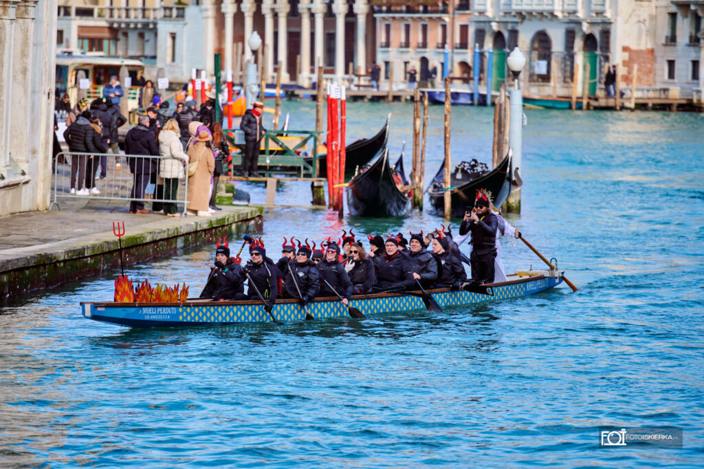 Długa, wąska niebieska łódź sportowa udekorowana motywem ognia na dziobie. Cała załoga jest ubrana na czarno i ma na głowach opaski z czerwonymi rogami diabła. Wioślarze synchronicznie zanurzają wiosła w wodzie.