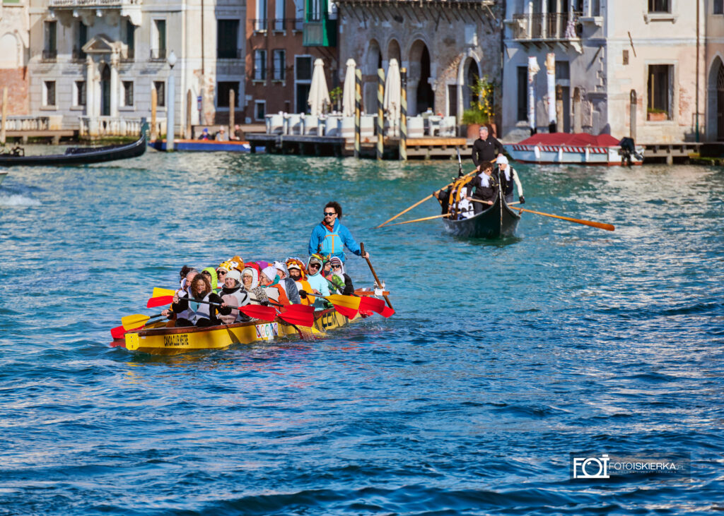 Żółta łódź wypełniona ludźmi w kolorowych, pluszowych kostiumach zwierząt (m.in. pandy, lwa i żaby). Na rufie stoi sternik w jaskrawoniebieskiej kurtce, kierujący łodzią za pomocą długiego wiosła.