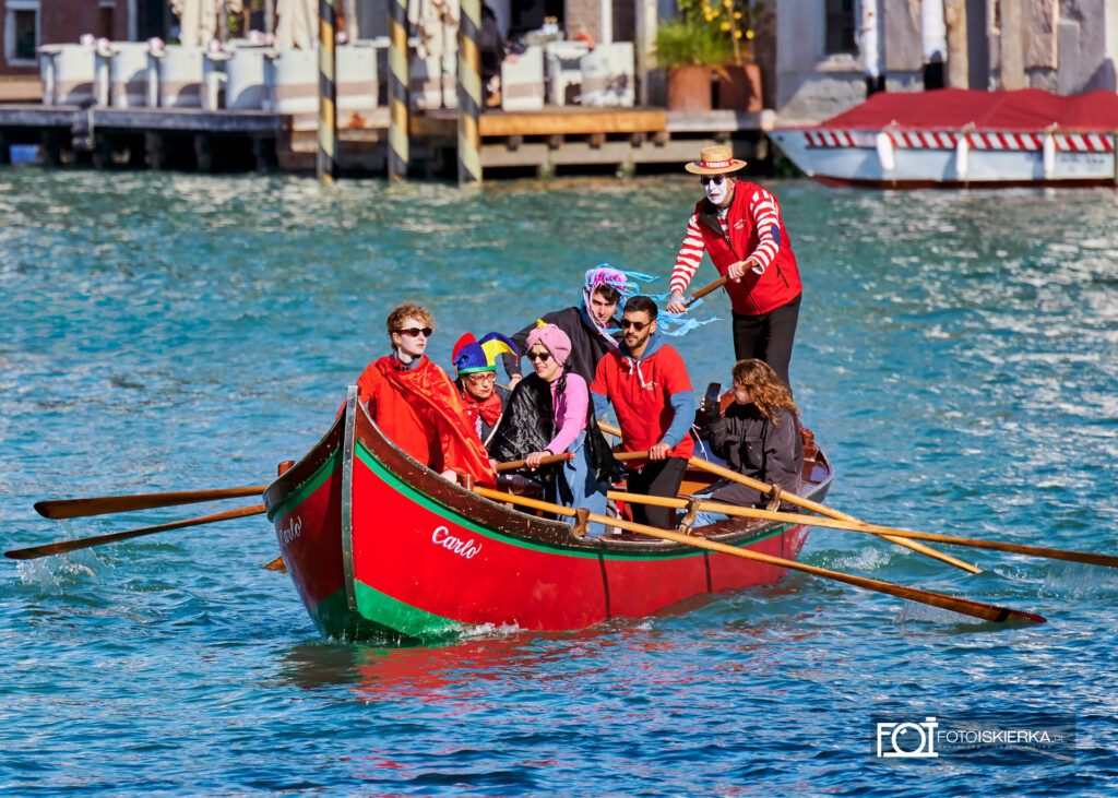Czerwona łódź „Carlo” z załogą w różnorodnych strojach. Na dziobie osoba w jaskrawoczerwonej pelerynie, a sternik na rufie ma pomalowaną na biało twarz mima, słomkowy kapelusz i koszulkę w czerwone pasy.