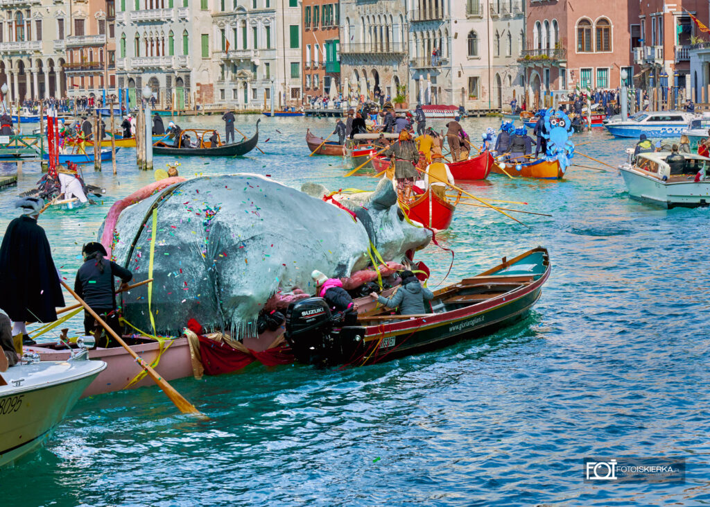 Wodna parada podczas Karnawału w Wenecji – widowiskowy korowód łodzi na Canal Grande. Uczestnicy w fantazyjnych kostiumach i maskach kultywują tradycję voga alla veneta. W oddali widoczna słynna Pantegana, symbolizująca ludowy charakter weneckiego święta Festa Veneziana sull’Acqua.