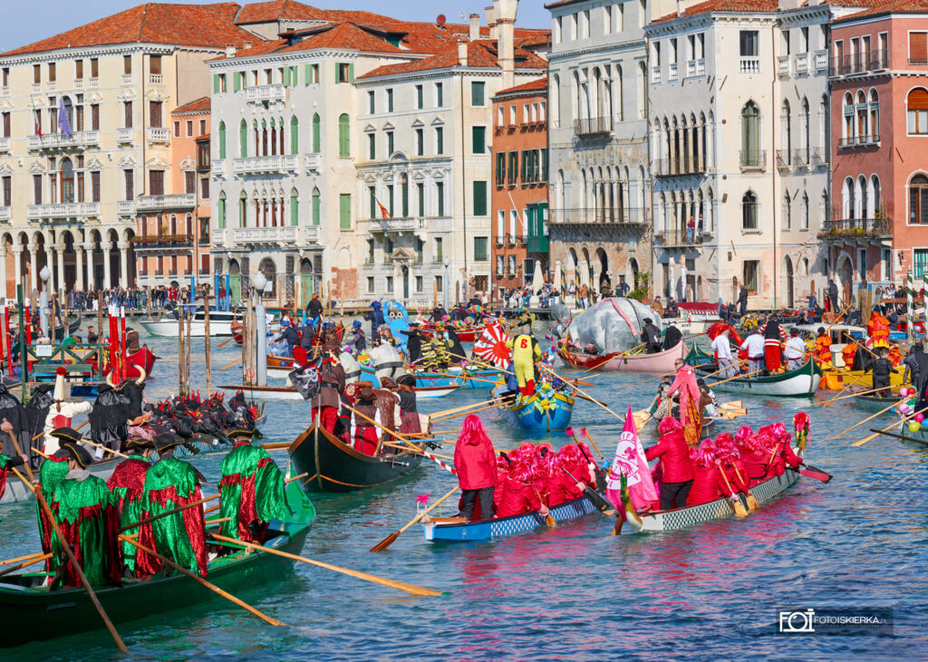 Wodna parada podczas Karnawału w Wenecji – widowiskowy korowód łodzi na Canal Grande. Uczestnicy w fantazyjnych kostiumach i maskach kultywują tradycję voga alla veneta. W oddali widoczna słynna Pantegana, symbolizująca ludowy charakter weneckiego święta Festa Veneziana sull’Acqua.