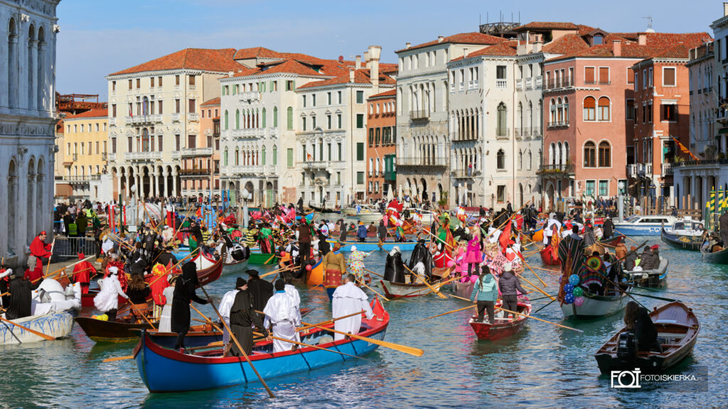 Parada wodna Festa Veneziana podczas Karnawału w Wenecji. Kolorowy korowód tradycyjnych łodzi na Canal Grande, w tym sandoli i caorline, płynący w stronę mostu Rialto. Widoczne radosne tłumy uczestników w maskach i kostiumach na tle historycznej zabudowy miasta.